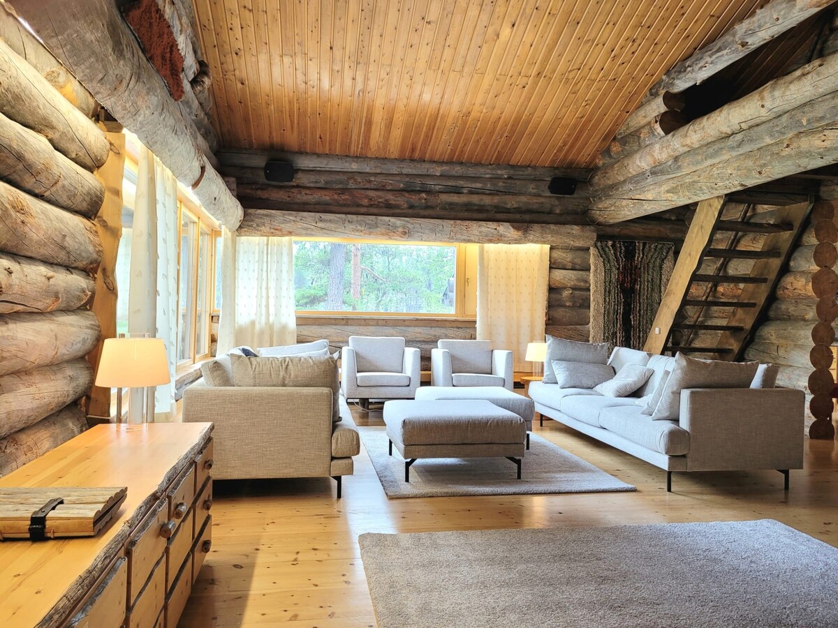 A spacious living area features a high wooden ceiling lined with logs, complemented by large windows allowing natural light. Comfortable seating includes a sectional sofa and armchairs arranged around a coffee table. A cozy area rug adds warmth to the wooden floor.