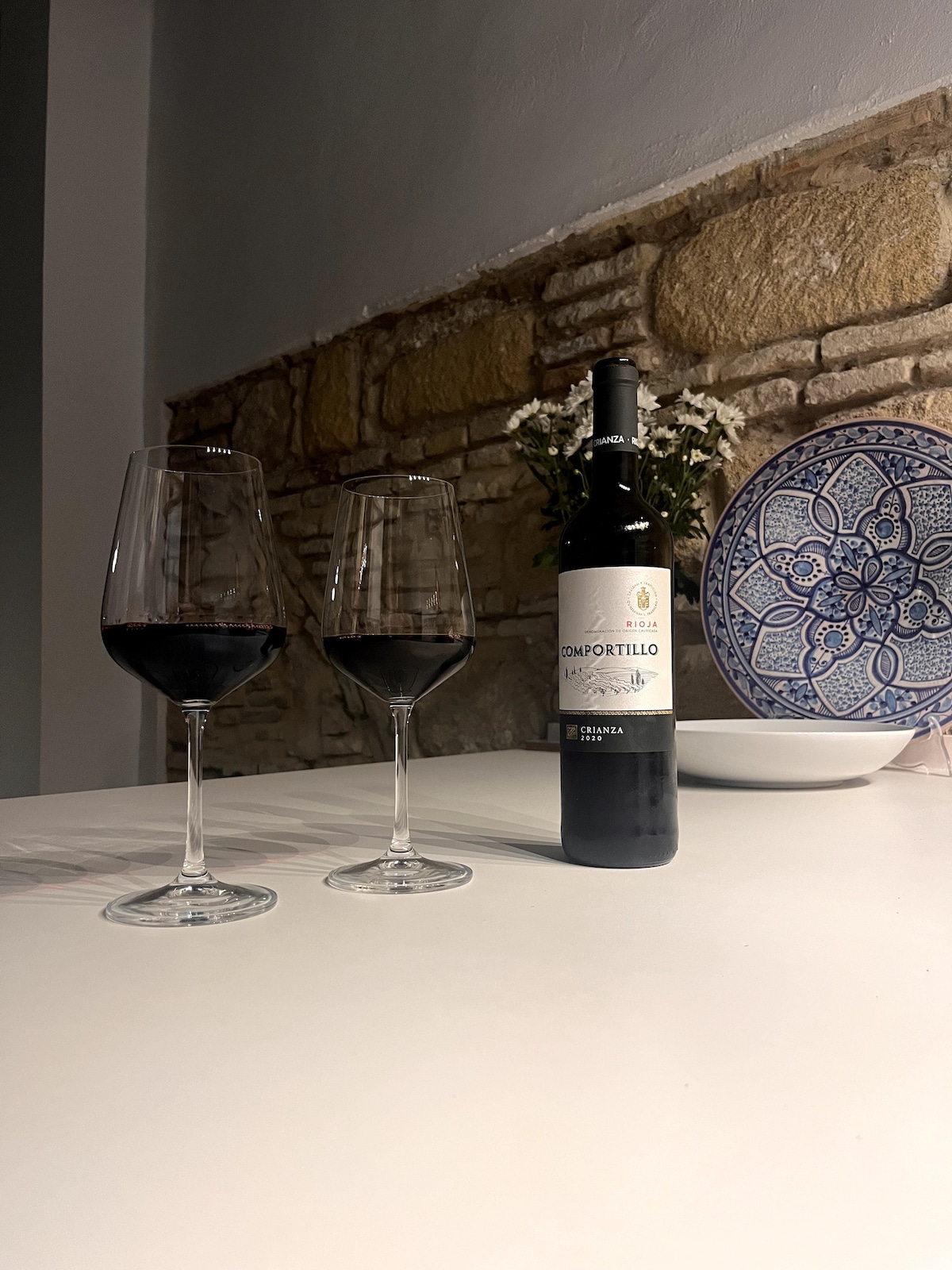 Two glasses of red wine are positioned elegantly on a white table, accompanied by a bottle of wine. In the background, an intricate blue and white decorative plate is visible, complemented by a stone wall that adds texture to the setting.