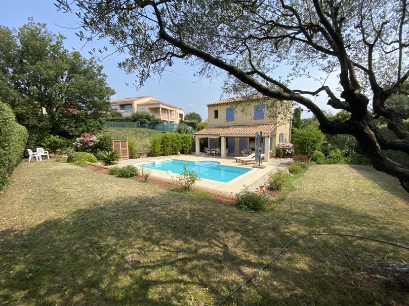 A view of the villa from the garden is presented, featuring a rectangular swimming pool surrounded by a well-maintained lawn. Tall trees and colorful shrubs border the area, providing a sense of privacy and tranquility. The two-story villa is partially visible in the background.