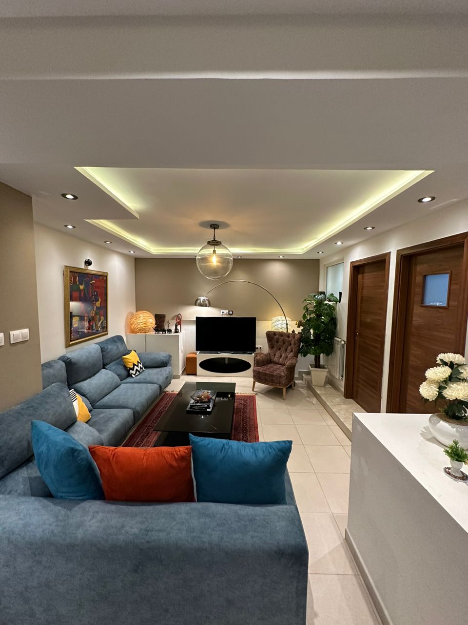 A spacious living area is highlighted by a cozy sectional sofa adorned with colorful cushions. A modern entertainment unit houses a large television. Soft lighting from the ceiling accents the room's design, while a decorative rug adds warmth. Two wooden doors lead to adjoining spaces.