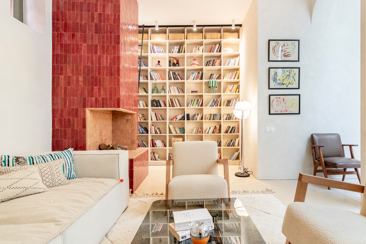 A serene sitting area features a neutral sofa and a cozy chair, with a bookshelf filled with various books as a backdrop. Soft lighting emanates from a floor lamp, while a modern coffee table holds decorative items and books, creating a welcoming space.