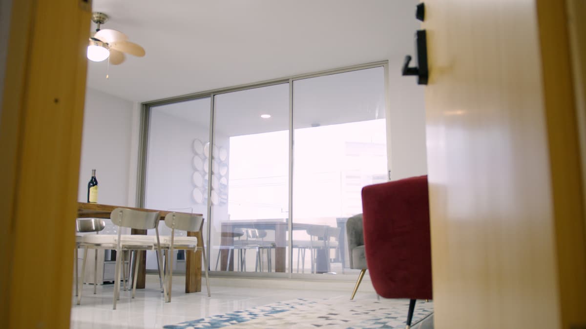 The entrance leads into a well-lit open space featuring large glass sliding doors that reveal a bright dining area. A wooden table with light-colored chairs is visible alongside a plush red armchair, enhancing the welcoming ambiance.