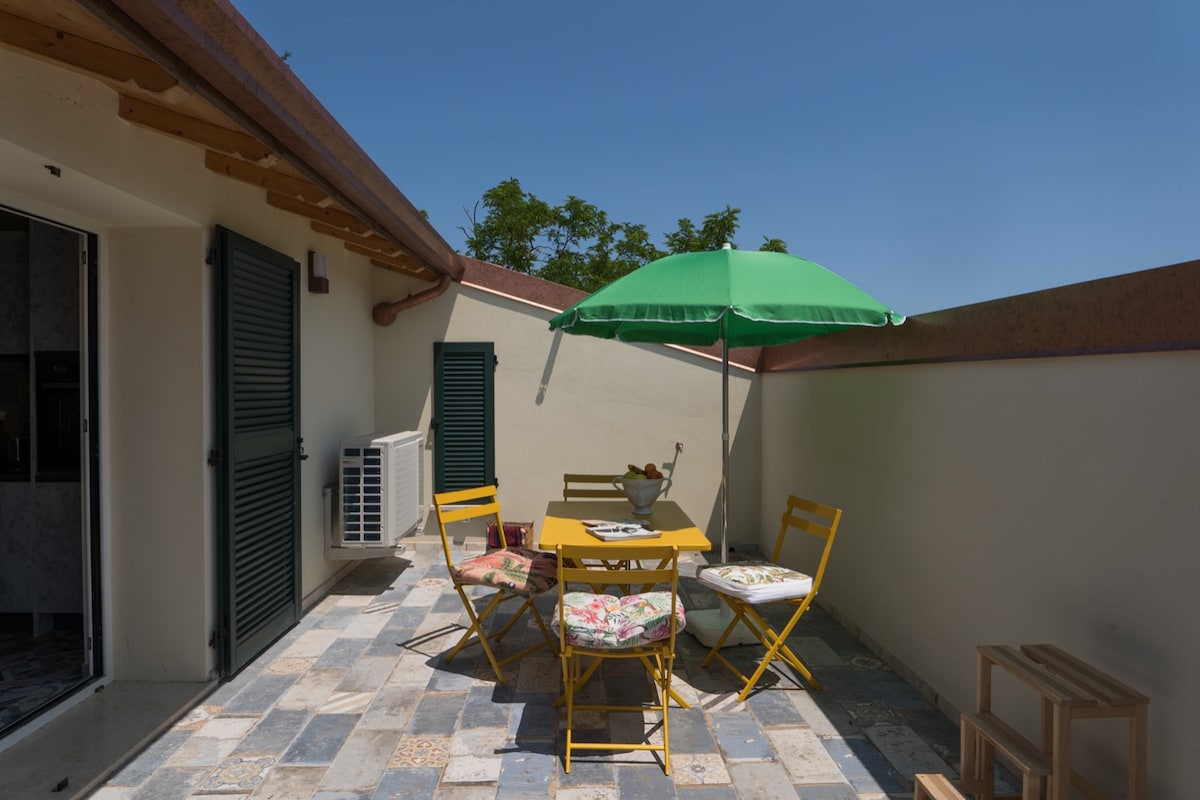 An outdoor terrace features a yellow dining table surrounded by four matching chairs, adorned with floral cushions. A green umbrella provides shade over the table, while a stone floor extends across the space. The surrounding walls add privacy, contributing to a cozy atmosphere.
