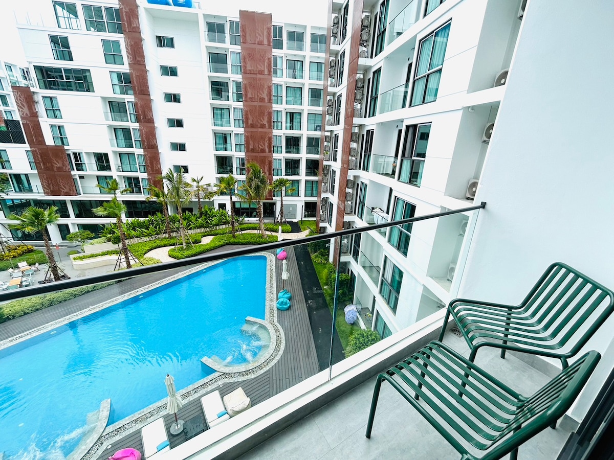 A balcony view captures the swimming pool bordered by modern landscaping. Two green metal chairs are positioned on the glass-railed balcony, providing a relaxing space to enjoy the outdoor scenery. The pool area features lounge chairs and a tranquil atmosphere.