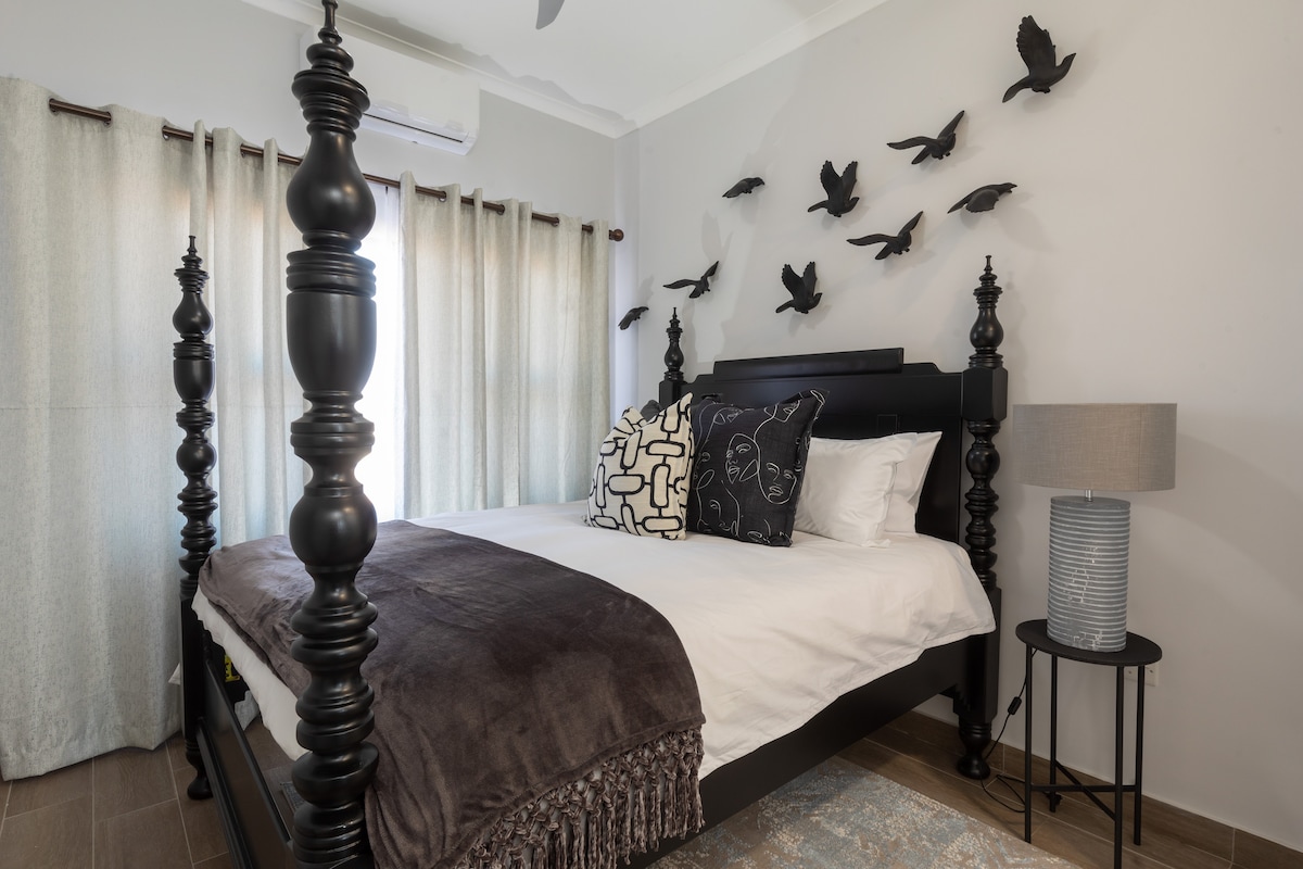 A well-appointed bedroom features a spacious bed adorned with decorative pillows and a soft throw. Black carved posts frame the bed, while a bedside lamp and a textured vase add subtle decor. Natural light enters through floor-length curtains, enhancing the room's serene ambiance.