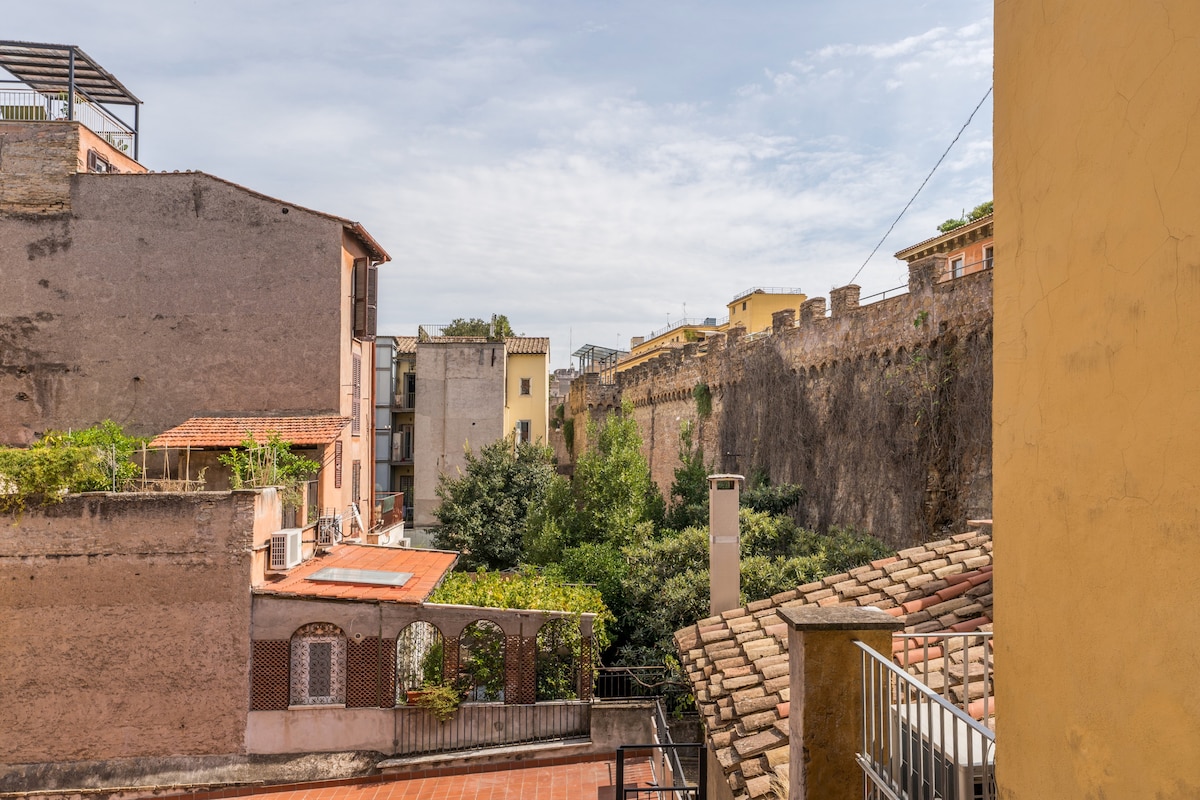 A view of the surrounding buildings and cityscape is presented, featuring quaint architecture and a historic stone wall in the background. Lush greenery is visible in the foreground, adding a touch of nature to the urban setting. The sky is bright and slightly cloudy.
