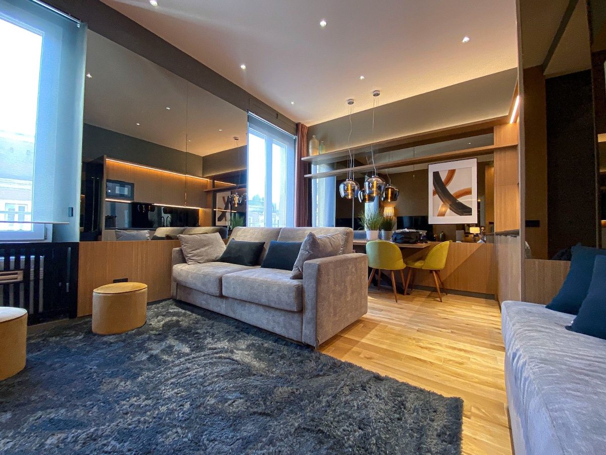 A modern living area features a plush, light-colored sofa and a large area rug with a soft texture. There are wooden accents throughout, with a dining area visible in the background, highlighted by stylish lighting and a contemporary art piece.