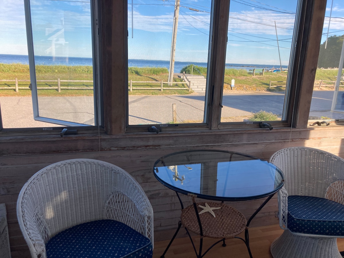 A sunlit enclosed front porch features large windows offering views of the beach and ocean. Two wicker chairs with blue cushions are positioned around a glass-topped table, creating a cozy space for relaxing while enjoying the ocean breeze.