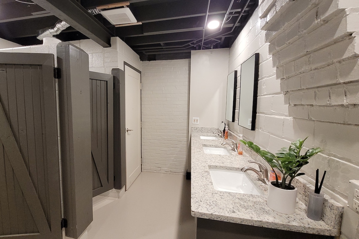A communal restroom facility features multiple stalls with wooden doors and two sinks positioned against a sleek granite countertop. Mirrors are mounted above the sinks, and a small potted plant adds a natural touch to the space. The walls are painted in a soft, neutral tone.
