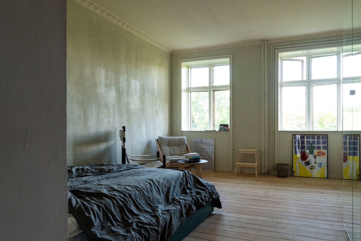 A spacious bedroom features large windows allowing natural light to fill the area. A simple bed is placed alongside a comfortable armchair and a small table. The wooden floor adds warmth, while colorful artwork is visible in the corner, enhancing the room's atmosphere.