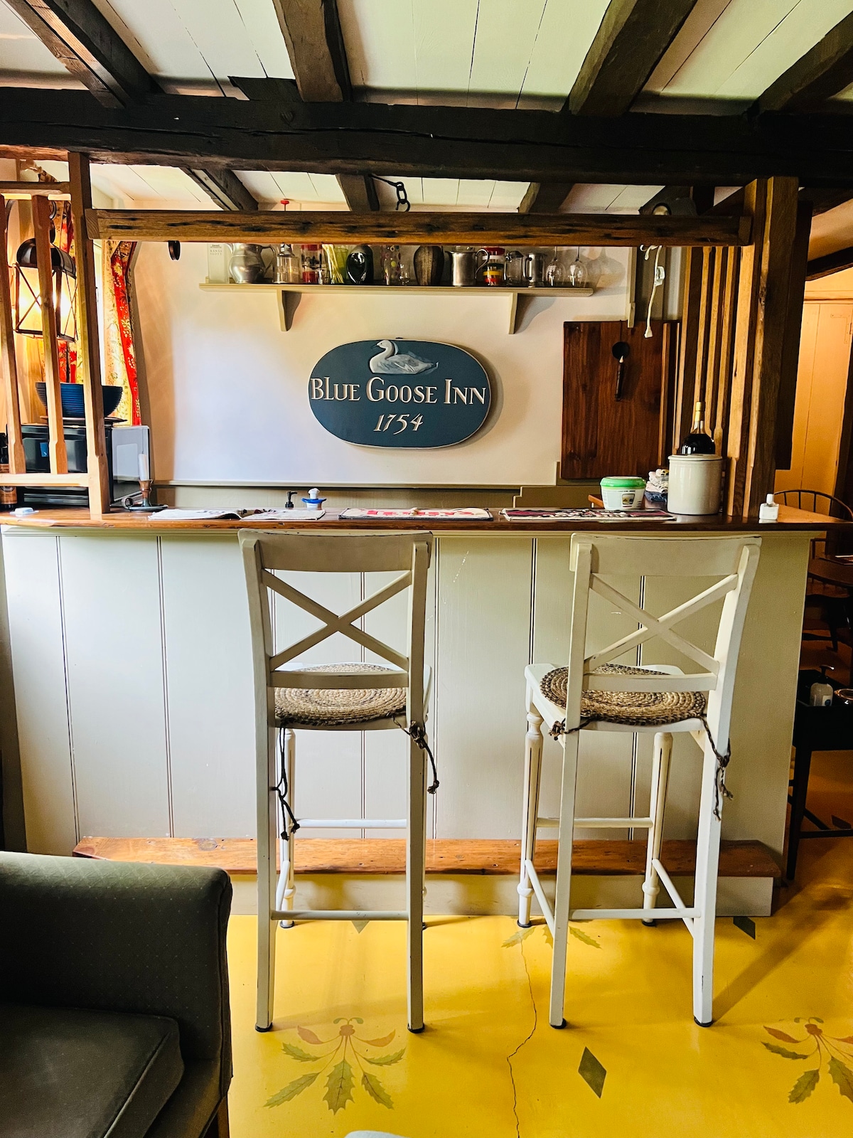 A quaint kitchenette area features a light-colored bar with two high wooden stools. Shelves above hold glass jars and kitchen essentials, while a sign labeled 'Blue Goose Inn 1754' hangs prominently on the wall. Bright yellow flooring complements the warm atmosphere.