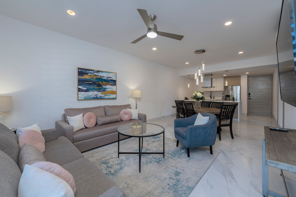 A modern living area features a spacious sofa and a cozy accent chair, complemented by a glass coffee table. Tasteful artwork adorns the wall, while natural light brightens the room. An open layout connects to the dining area and kitchen in the background.