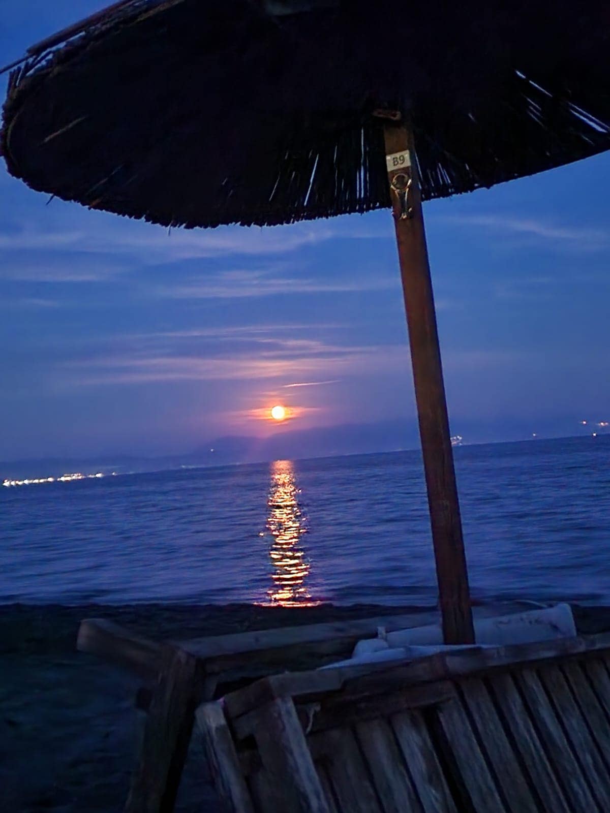A straw beach umbrella is positioned near the shoreline, with the calming sea visible beyond. A sunset casts a warm glow, reflecting on the water's surface. Distant lights from the coastline twinkle softly against the twilight sky.