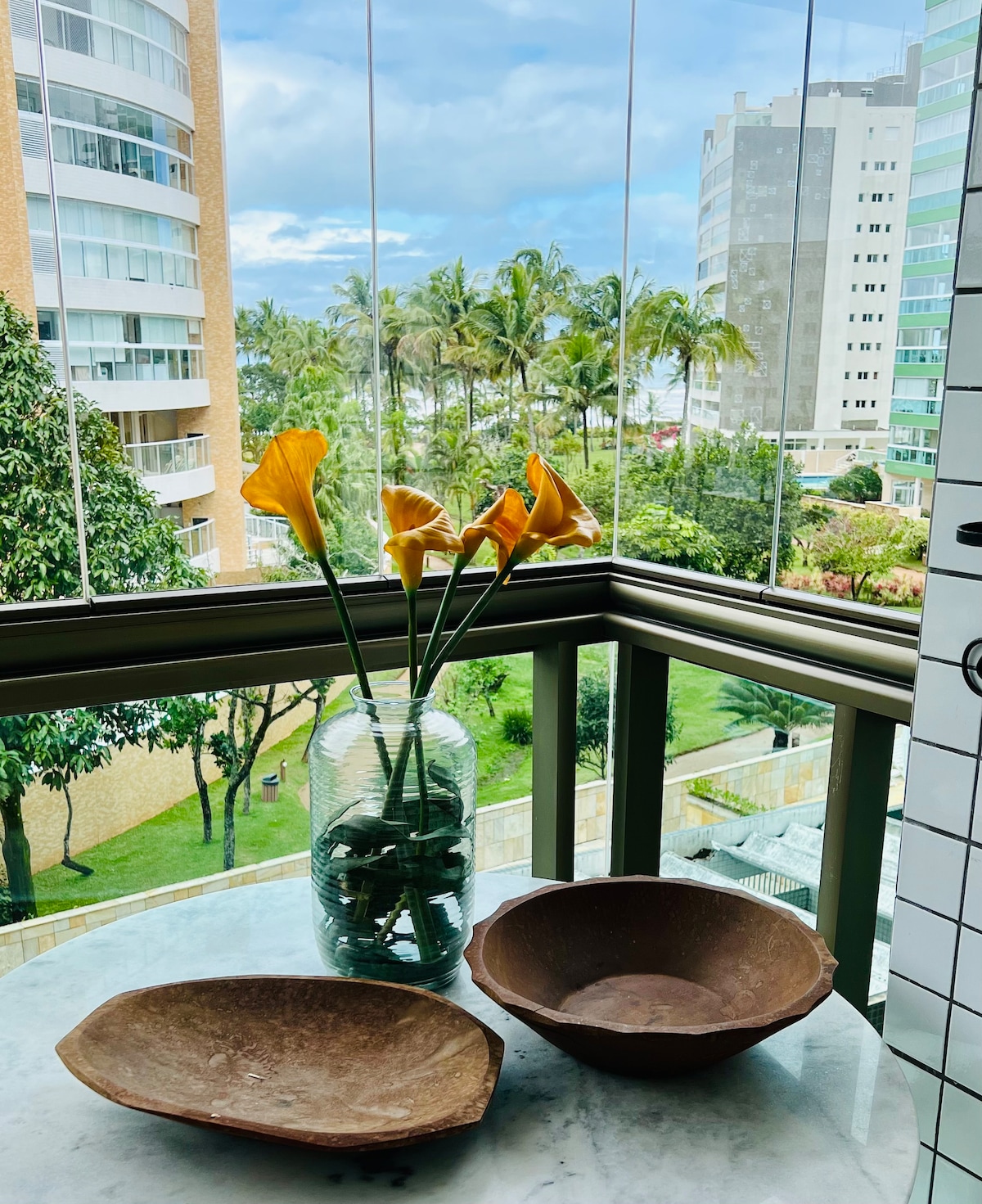 A glass vase filled with yellow calla lilies is positioned on a marble table. Two shallow bowls, one made of wood and the other ceramic, rest beside the vase. In the background, lush greenery and tall buildings can be seen through large windows.