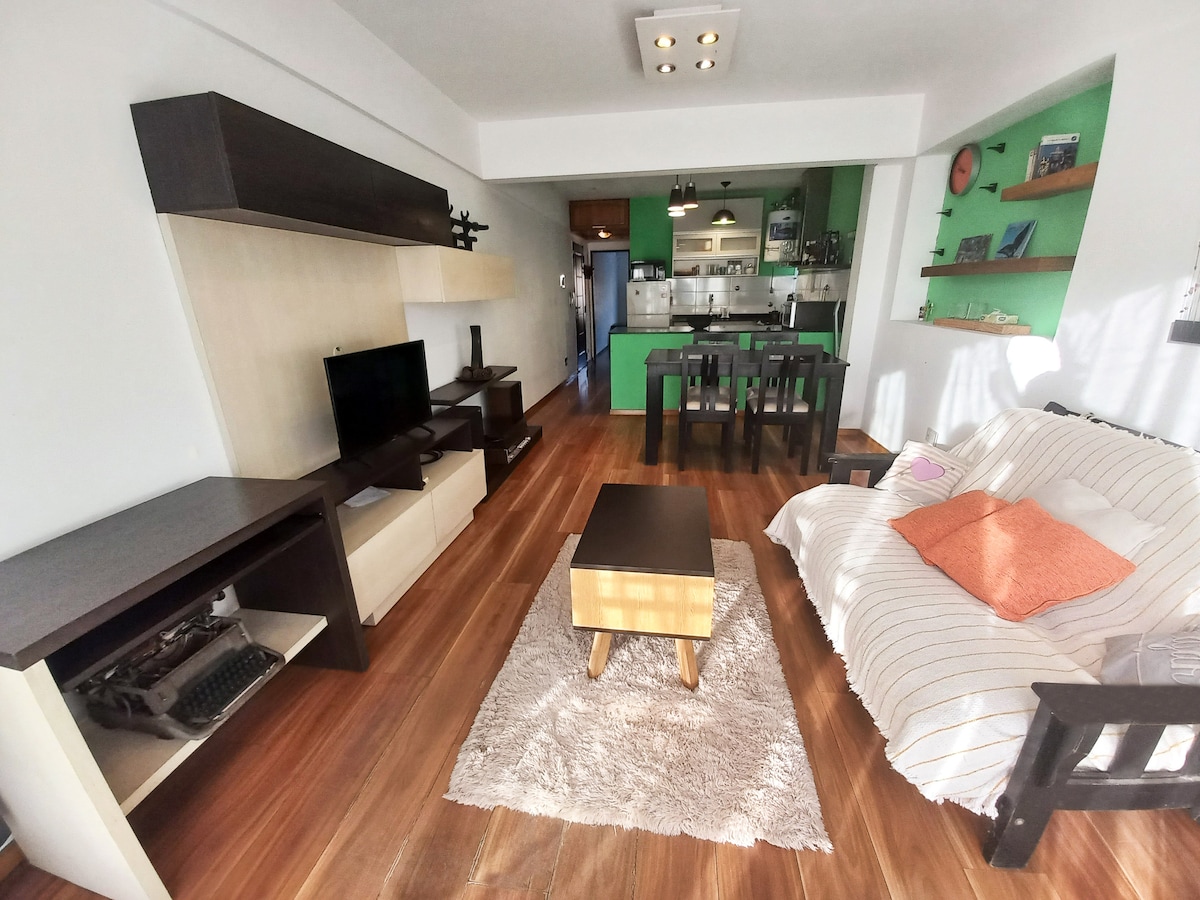 A bright living area features a minimalist design with a dark wooden shelf and entertainment unit. A light-colored sofa and a small coffee table sit on a textured area rug. The green kitchen area is visible in the background, complemented by a dining table with chairs.
