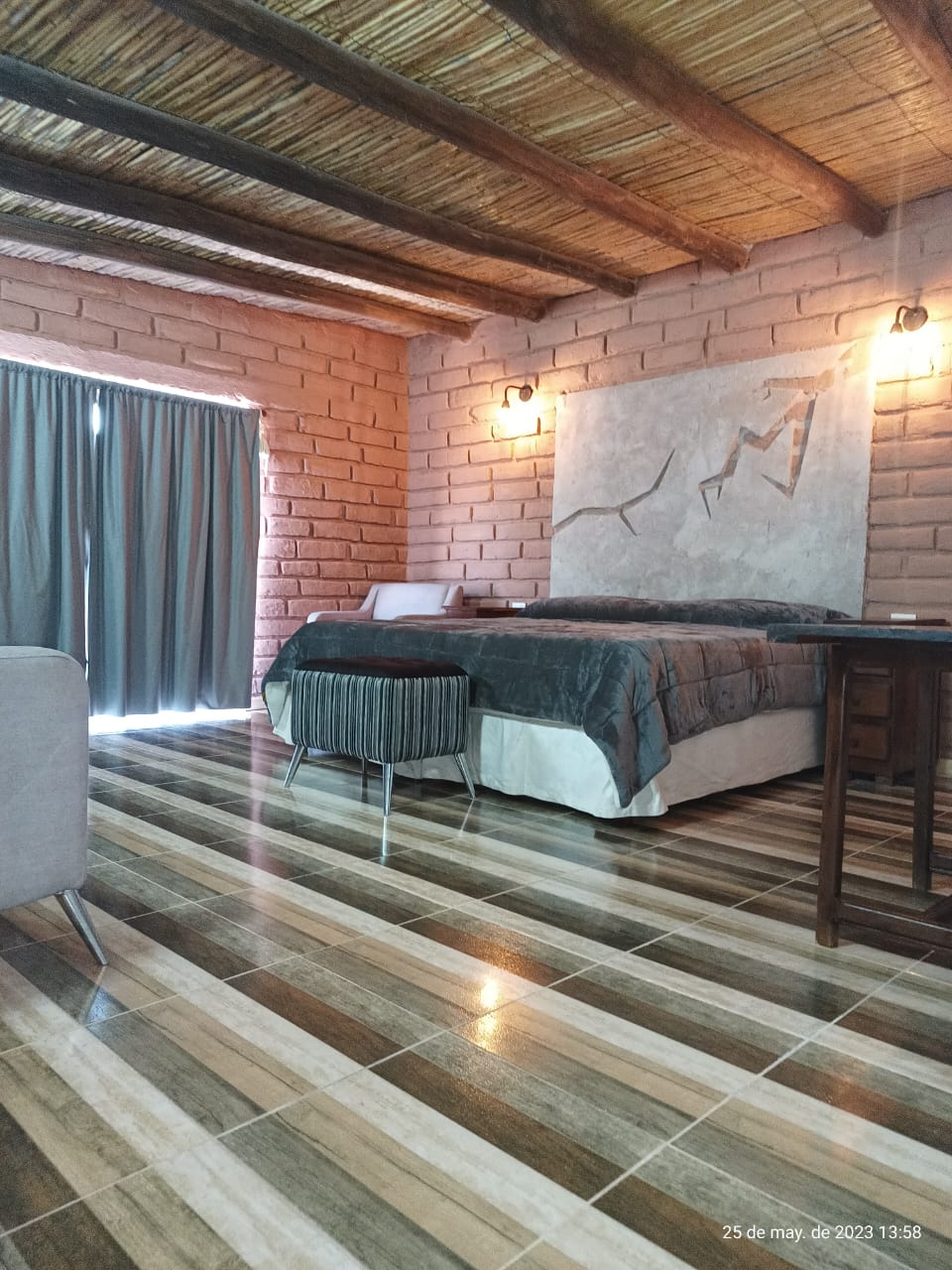 A cozy suite features textured walls and a spacious bed, adorned with soft bedding. A dark curtain partially filters natural light from large windows, accentuating the earthy tones of the floor tiles. A rustic table complements the inviting atmosphere.