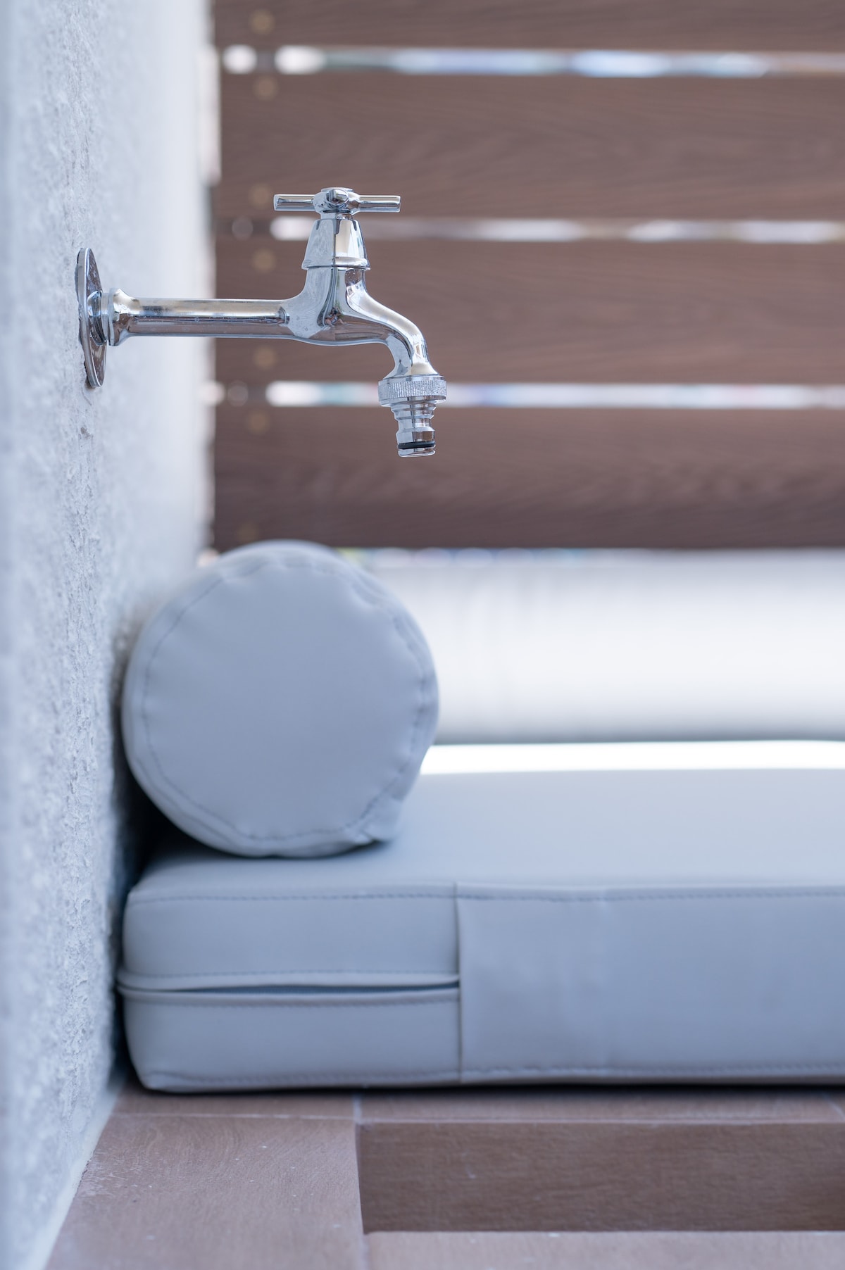 A sleek chrome faucet is mounted on a textured wall, positioned above a comfortable gray cushion. The cushion is part of a cozy seating area, creating an inviting space for relaxation. Soft natural light enhances the modern aesthetic.