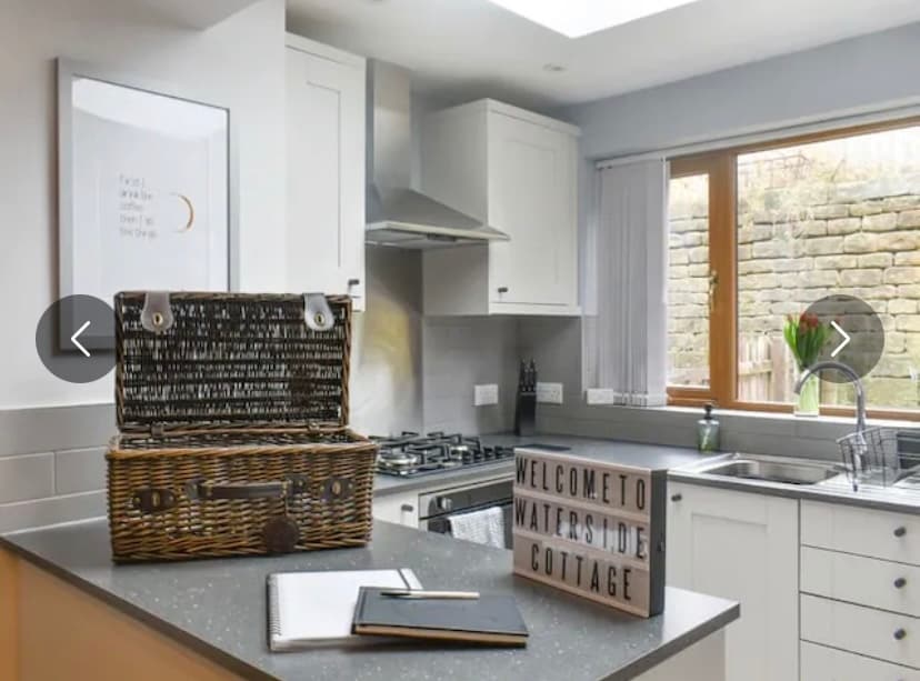 The kitchen features modern appliances and a neutral colour scheme. A wicker basket sits atop the grey countertop, accompanied by a welcome sign and a notebook. Natural light enters through the window, highlighting the uncluttered, functional design.