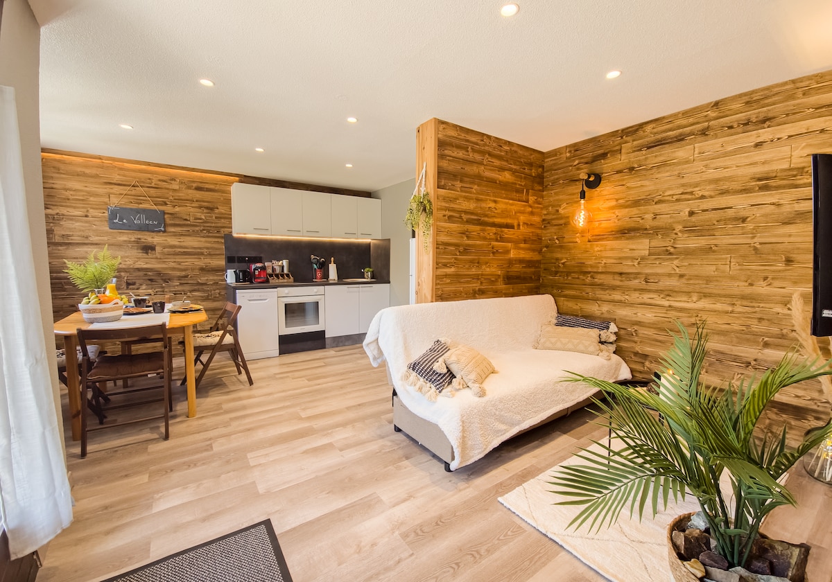 A modern living area combines a cozy sofa with a neutral throw and a wooden accent wall. Adjacent, a dining table is seen set for meals, with a variety of kitchen appliances and decor visible in the background.