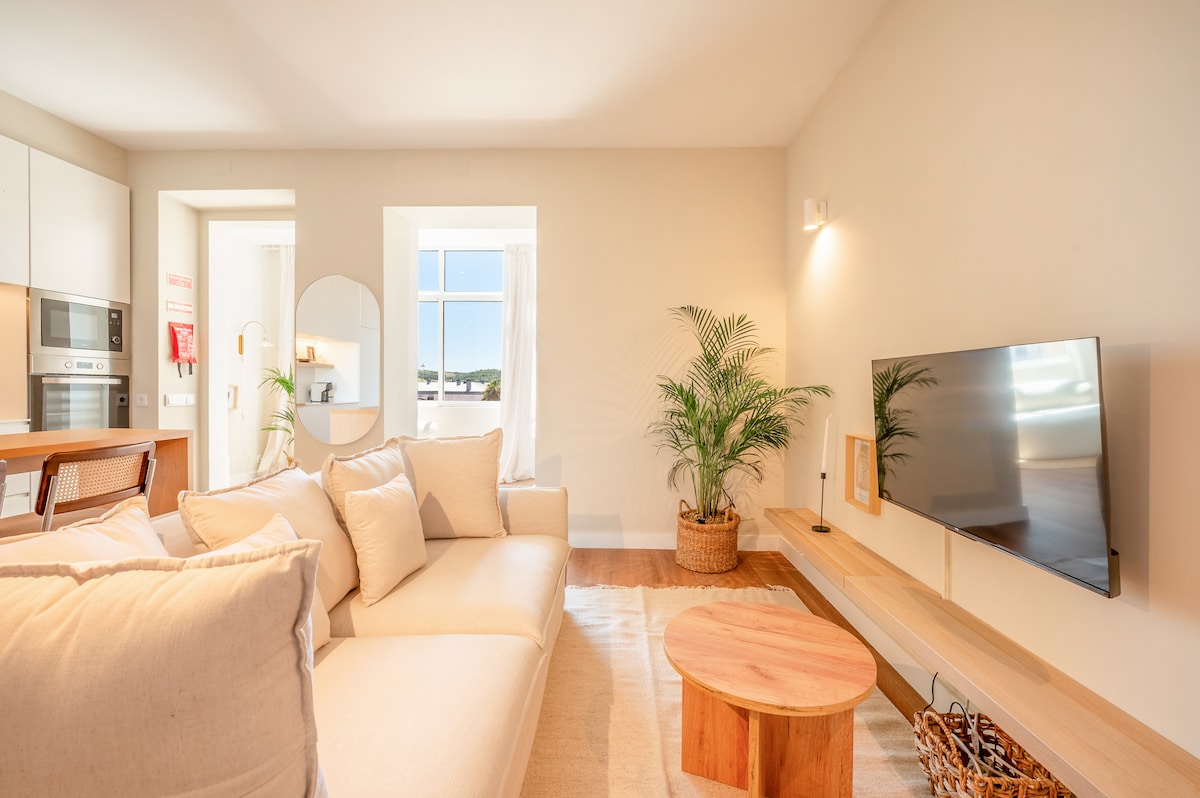 A well-lit living area features a spacious sectional sofa adorned with neutral cushions. A wooden coffee table sits in front, complemented by a decorative plant. A wall-mounted television is visible, alongside a simple shelf holding framed images. Large windows allow natural light to brighten the space.