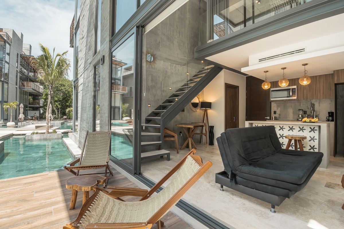 A modern living space is highlighted by an open layout, featuring a sleek black sofa and a wooden dining table. Large glass doors offer a view of the swim-up pool. Sun loungers are positioned outside, surrounded by lush greenery and natural light.