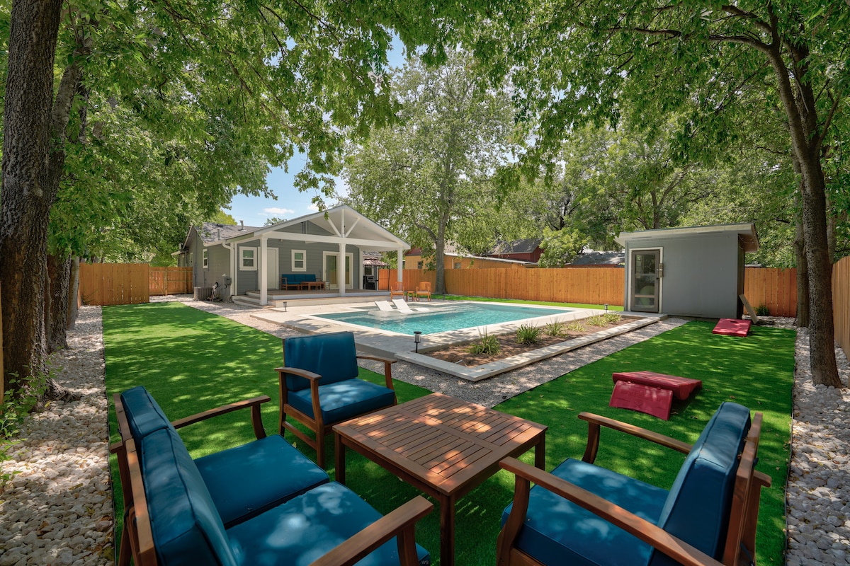 An outdoor seating area is presented with blue chairs arranged around a wooden table. The space features a well-maintained lawn, a heated pool, and a covered patio in the background. Surrounding trees provide shade, creating a private and inviting atmosphere.