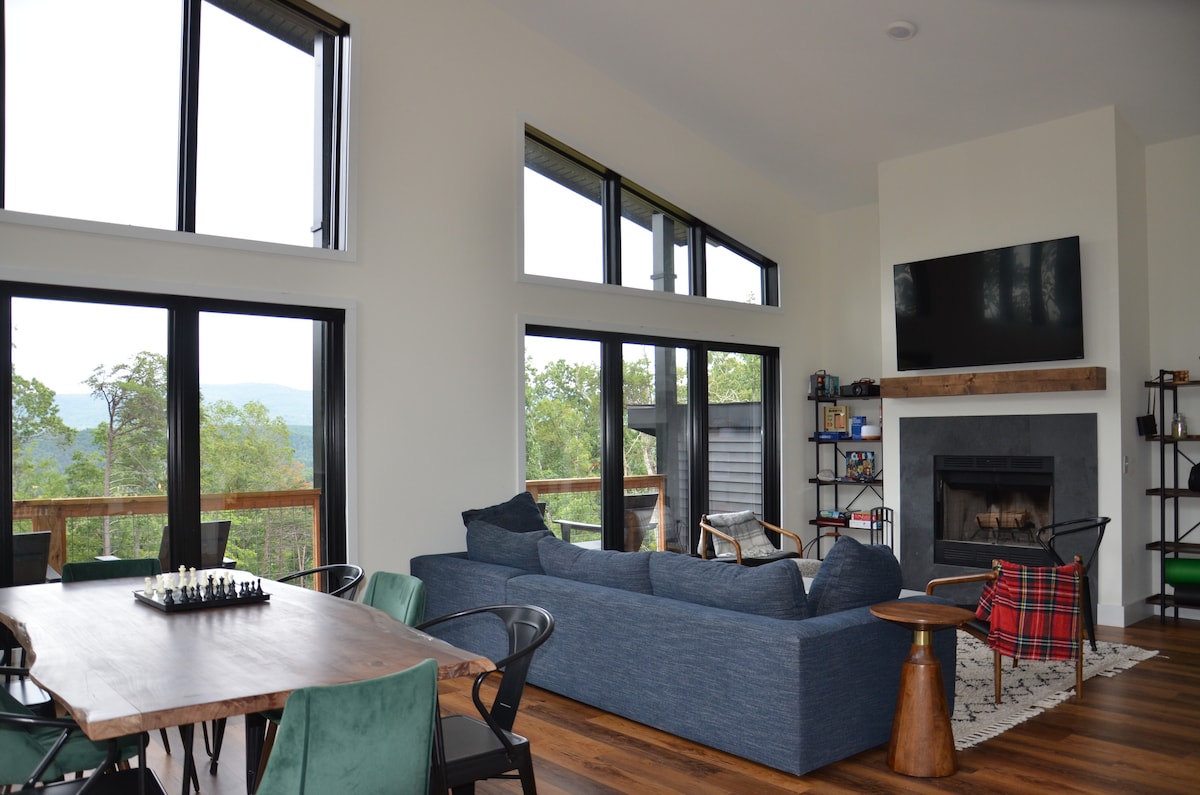 A spacious living area features large windows that frame views of the surrounding mountains and forest. A comfortable couch is positioned near a wood-burning fireplace, with a wooden coffee table and a chess set enhancing the inviting atmosphere. A dining table with green chairs completes the setup.