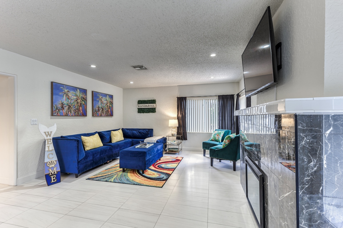 A spacious living room features a large blue sofa and a cozy green armchair. Colorful artwork decorates the walls, and natural light enters through the window. A flat-screen TV is mounted on the wall, and a patterned rug adds texture to the tiled floor.
