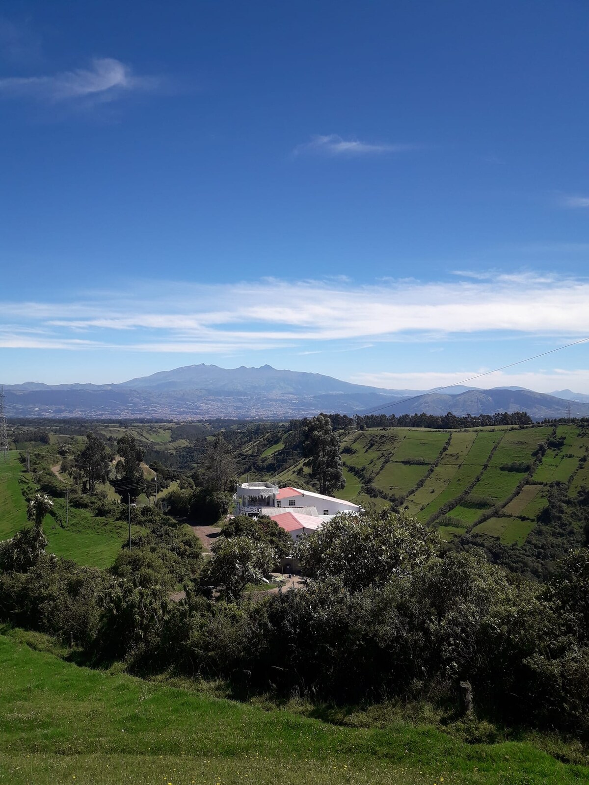 A scenic view captures a white building nestled among lush green hills, with expansive fields leading down the slope. The backdrop features distant mountains under a clear blue sky, while gentle clouds add depth to the landscape.