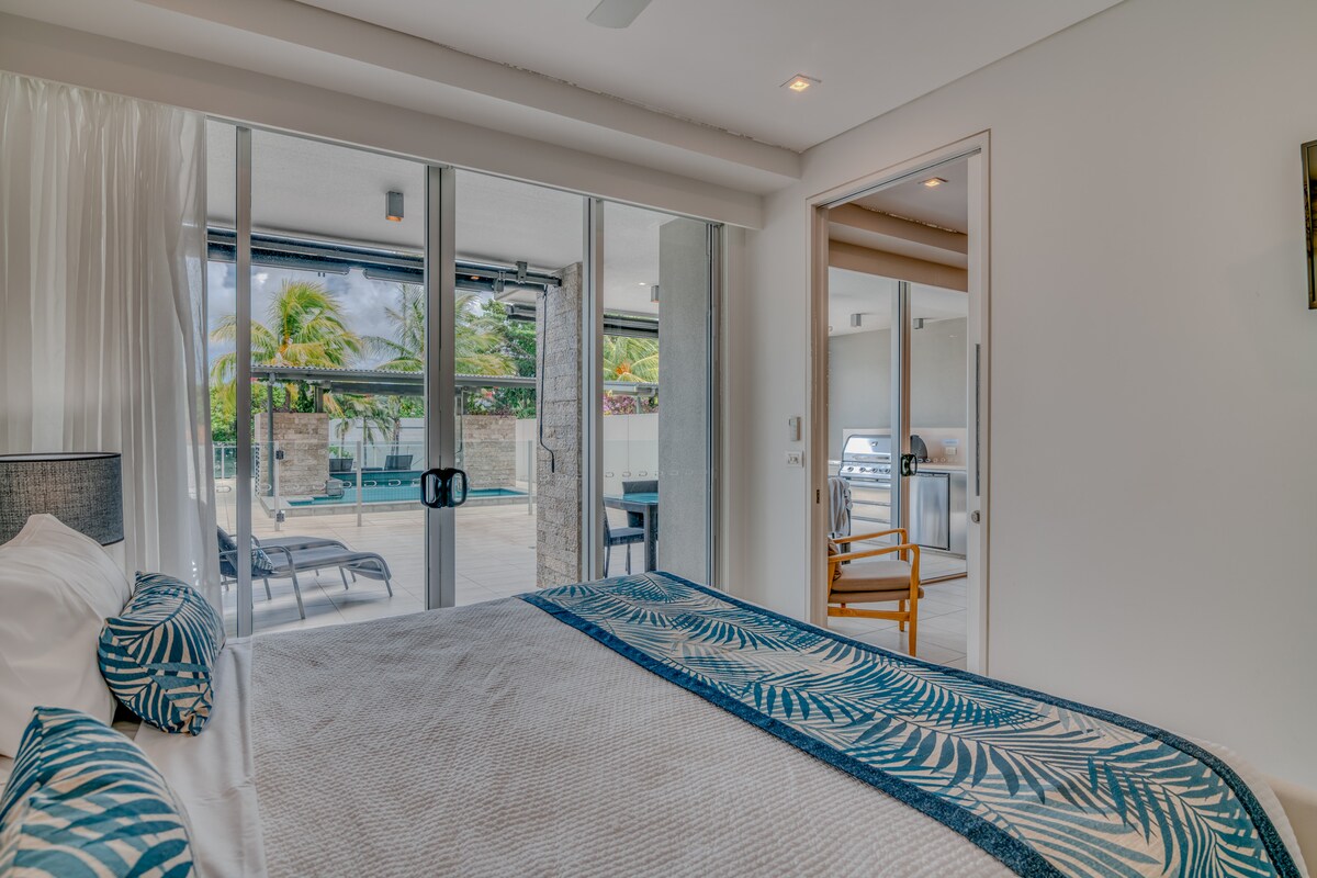 A spacious bedroom features a comfortable bed adorned with a patterned blanket. Large glass sliding doors provide access to a terrace area with outdoor seating and a barbecue. Natural light filters in, highlighting the casual decor and inviting atmosphere.