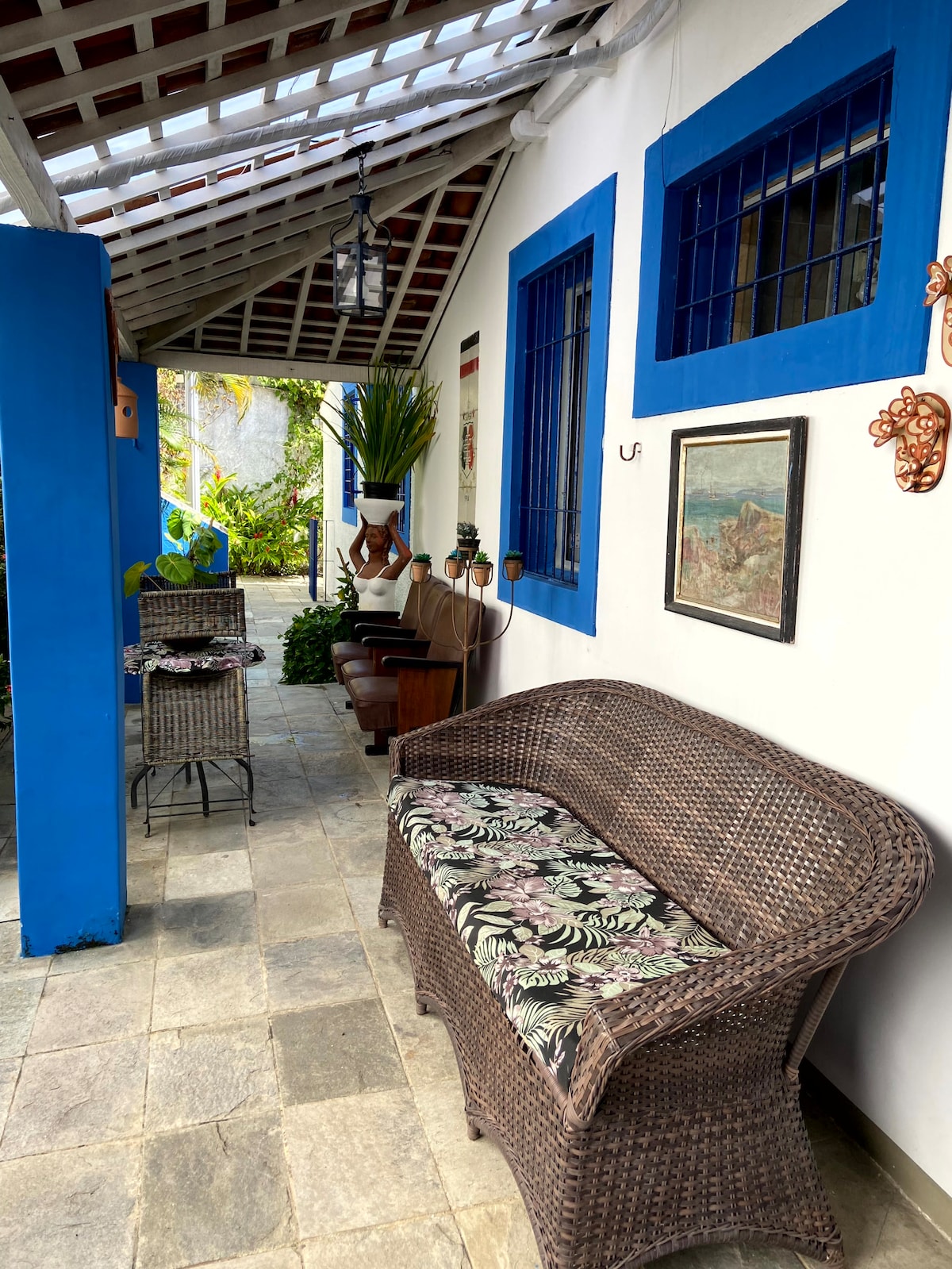 A welcoming corridor leads to an outdoor seating area, featuring a woven sofa adorned with tropical-patterned cushions. Decorative plants are positioned along the walls, while stone tiles provide a sturdy floor. Blue window frames add a pop of color, enhancing the inviting environment.