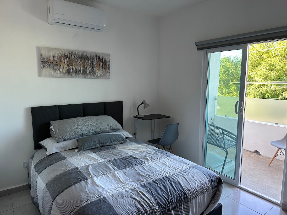 A spacious room features a comfortable bed dressed in neutral-toned linens and a textured throw. Natural light floods in from a sliding glass door that opens to a small balcony, complemented by a work desk and chair nearby.