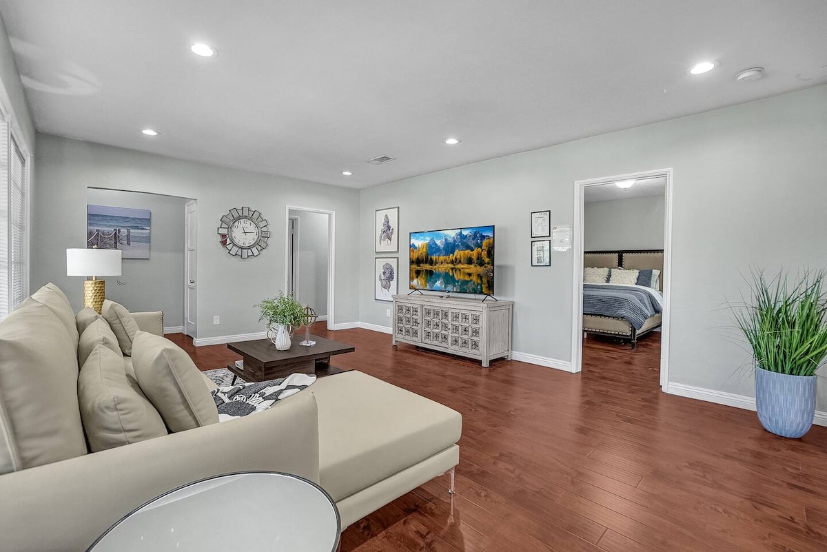 A bright and open living area contains a lavish sofa and a coffee table. A large 4K Smart TV is mounted on the wall, displaying a serene landscape. The space features warm wood flooring and leads to two additional rooms, enhancing the inviting environment.