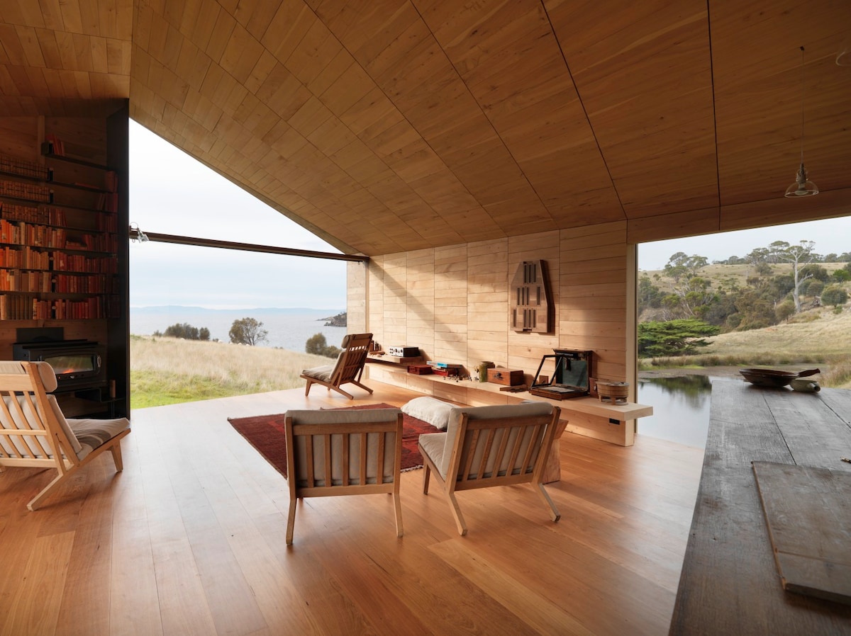 A spacious interior showcases natural wood finishes and large windows, allowing for panoramic views of the surrounding landscape. Comfortable seating is arranged thoughtfully, and a small bookshelf adds a touch of warmth to the space. Soft natural light fills the room, enhancing the tranquil atmosphere.