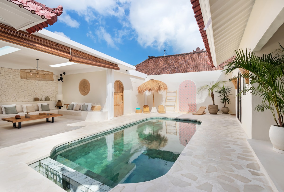 A Mediterranean-style outdoor area features a tranquil private pool surrounded by a textured stone deck. Sunbeds and a straw-roofed lounge area are positioned nearby, while decorative elements, including potted plants, enhance the inviting atmosphere. Natural light enhances the space, with a clear blue sky above.