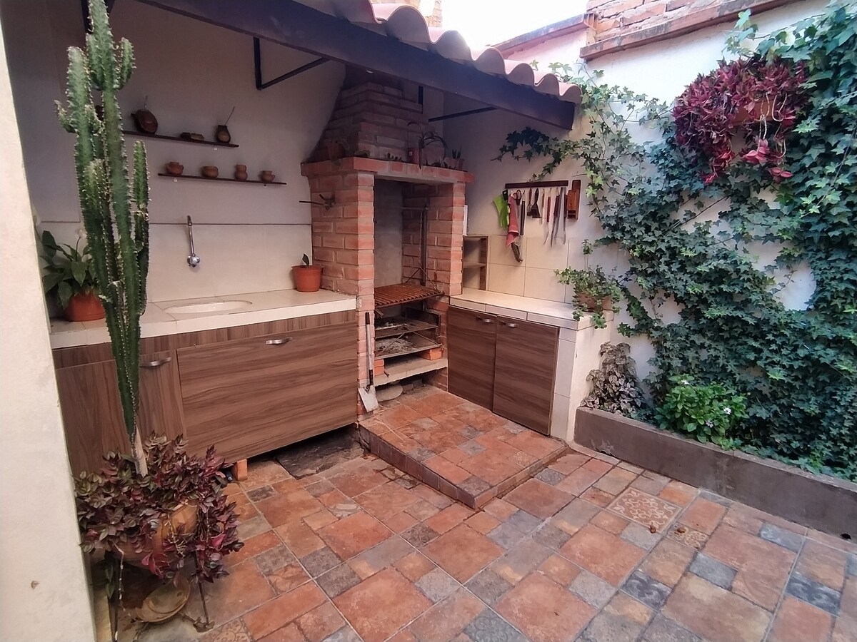 An outdoor kitchen area is presented, featuring a built-in brick barbecue and countertop space, surrounded by lush greenery. The floor is paved with patterned tiles, and planters filled with plants add a touch of nature to the environment.