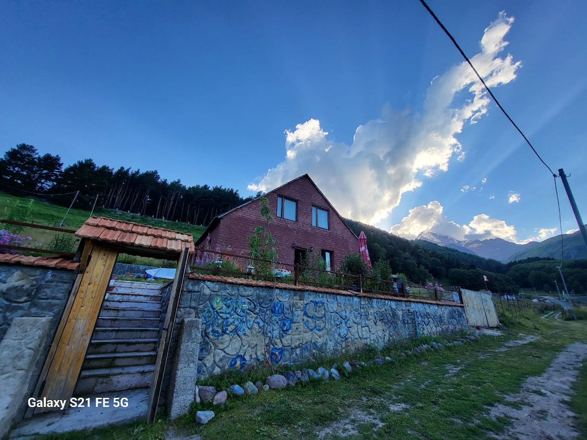 A red-brick guest house is framed by lush greenery, with a stone wall and artistic mural visible at the front. Majestic mountains rise in the background, under a dramatic sky filled with clouds and soft sunlight.
