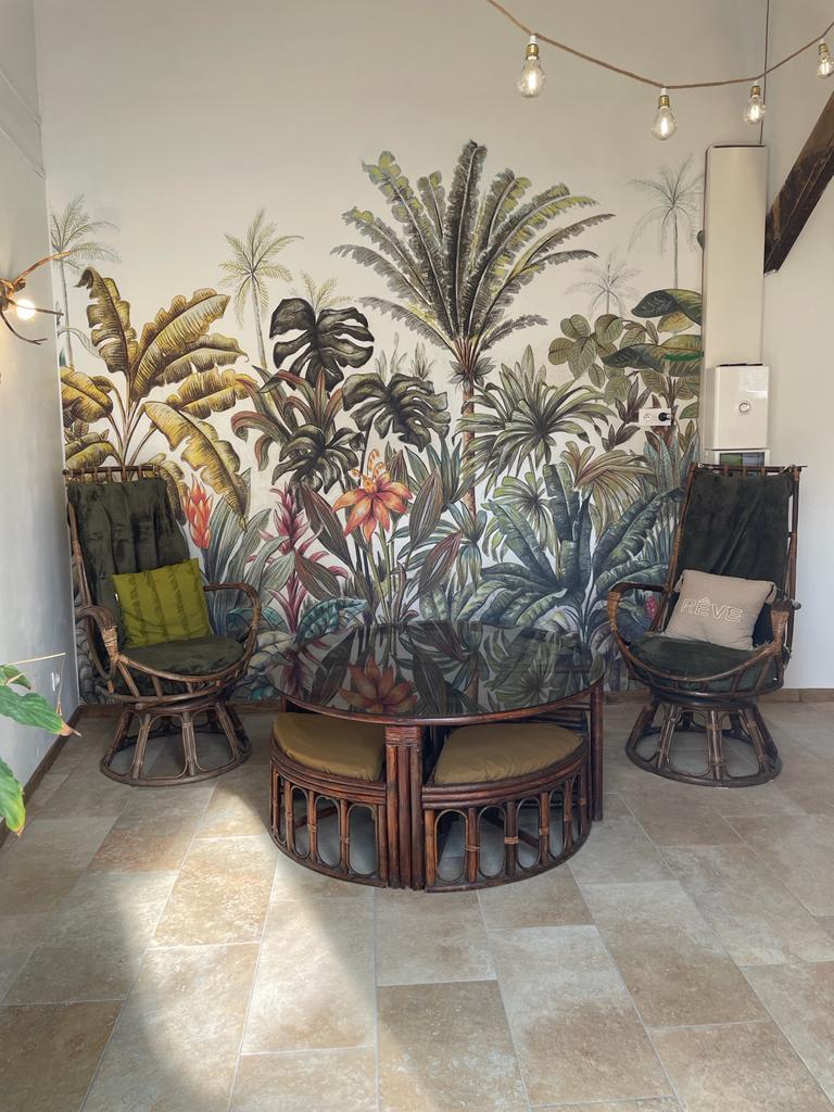 A cozy seating area features two rattan chairs with green cushions, positioned around a round glass table. A vibrant tropical mural of lush plants provides a lively backdrop, while warm string lights add a soft glow to the space.