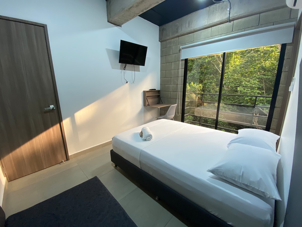 A simple bedroom presents a spacious bed with white linens and two pillows. A desk is positioned near a large window that offers views of greenery outside. A wall-mounted television is visible, and natural light filters through the glass, creating a bright and airy environment.