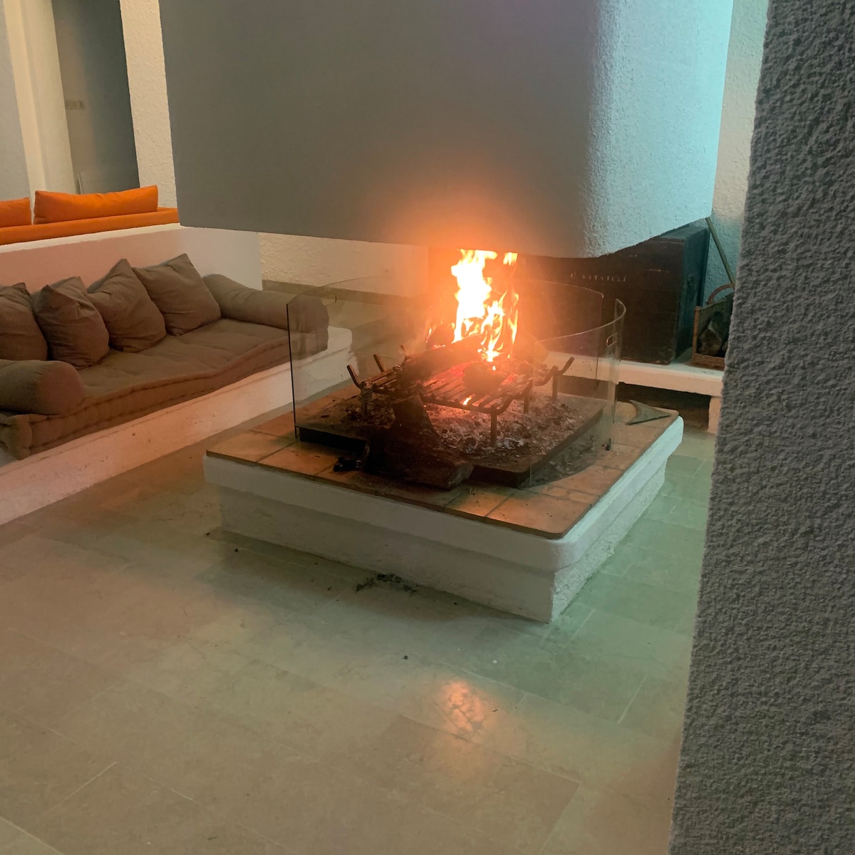 A modern fireplace is positioned in a spacious living area, featuring a glass barrier for safety. The flames create a warm ambiance, surrounded by a stone base. Comfortable seating is visible, inviting relaxation in this well-lit open space.