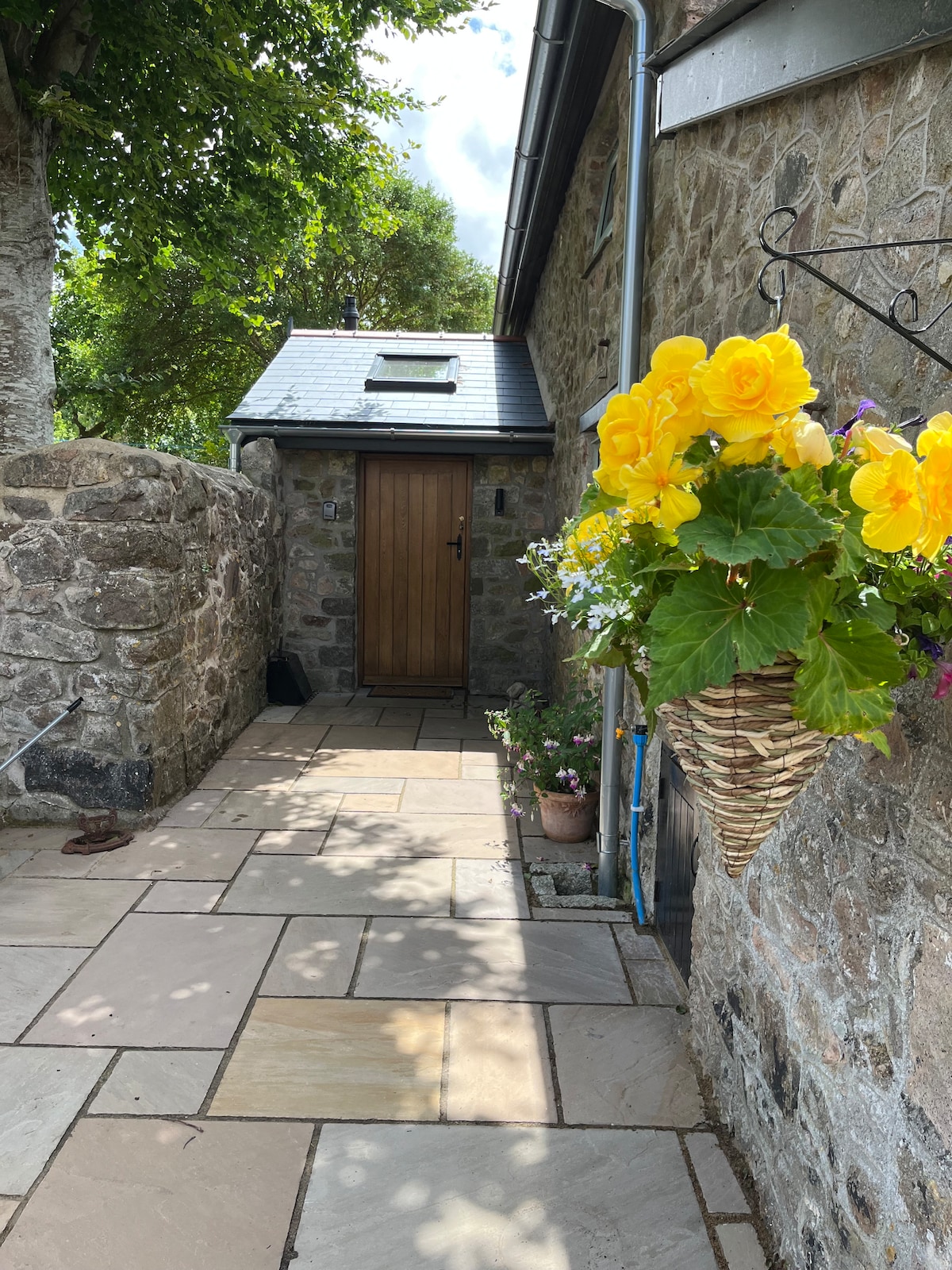 A rustic stone pathway leads towards a wooden door, framed by a lush hanging basket filled with bright yellow flowers. Natural light illuminates the scene, with green trees visible in the background, enhancing the inviting entrance of the cottage.