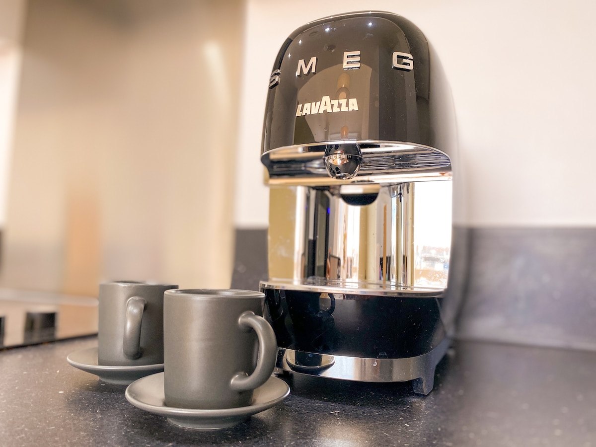 A sleek black coffee machine is displayed on a dark countertop, accompanied by two matching black ceramic cups and saucers. The machine's polished chrome finish reflects light, creating a modern and stylish appearance.