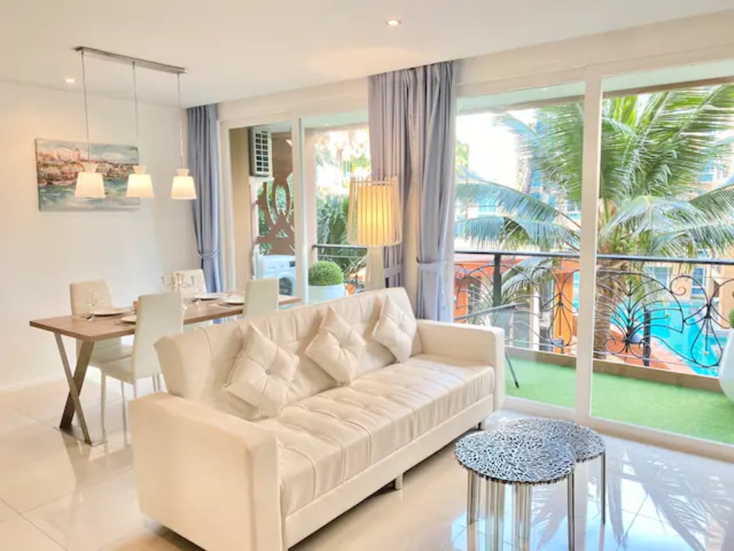 A light-filled living space features a plush white sofa adorned with decorative pillows. A glass table is set for dining, while large windows offer views of palm trees and a swimming pool. Soft pastel curtains add a touch of elegance to the atmosphere.