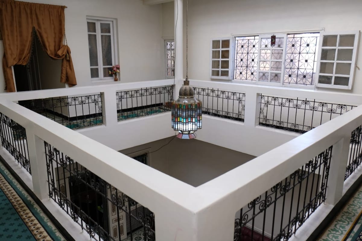A spacious interior courtyard is featured, surrounded by decorative iron railings and bright windows, allowing natural light to flow in. A traditional Moroccan lantern hangs from the ceiling, casting a warm glow throughout the area.