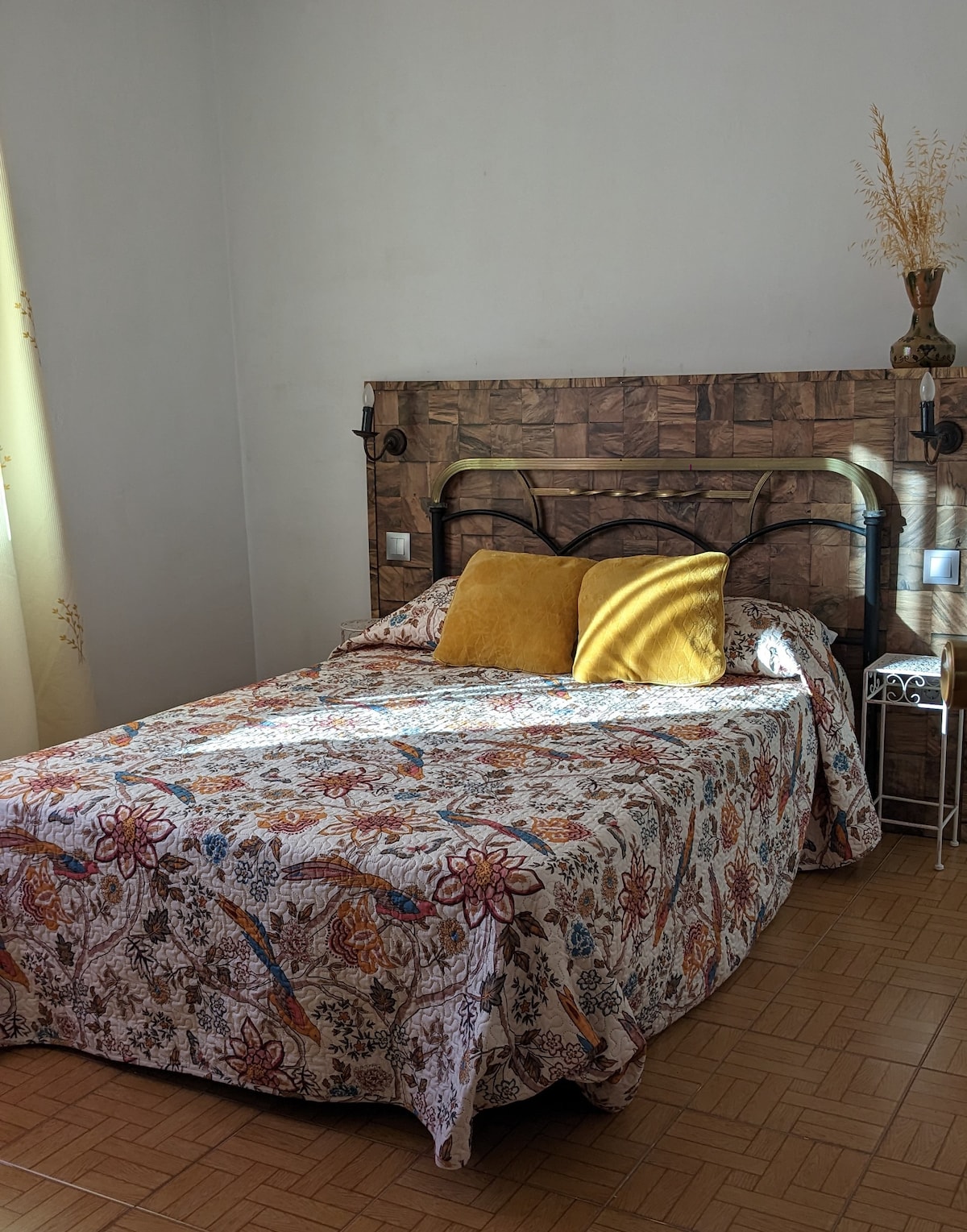 A cozy sleeping area is presented with a patterned bedspread featuring floral designs. Decorative pillows in warm tones are placed against a wooden headboard. A small bedside table holds a vase, and natural light filters through soft curtains, creating a serene atmosphere.