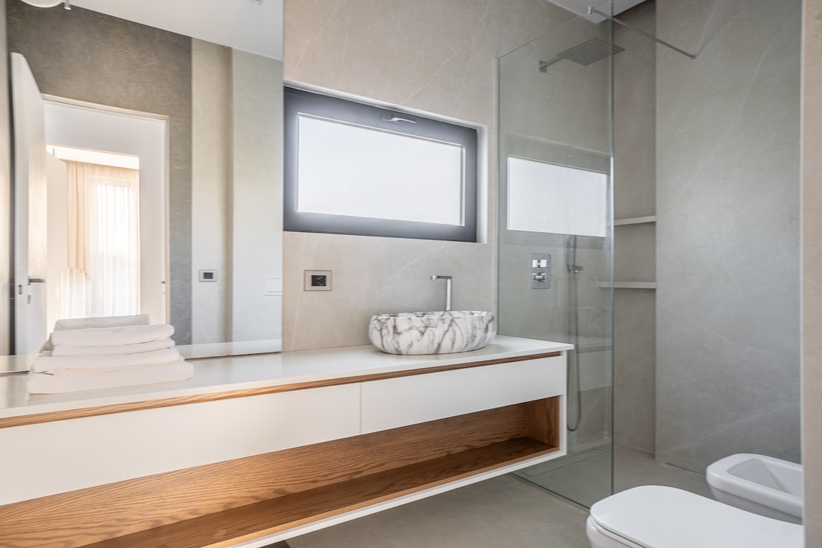 A modern bathroom features sleek stone walls and a minimalist design. A stylish sink with a marble basin is set atop a light wooden countertop. A spacious walk-in shower is enclosed by glass, and natural light enters through the frosted window.