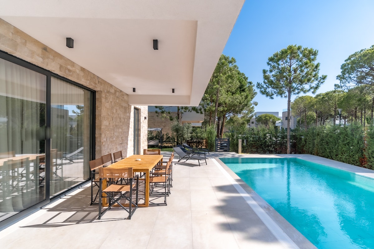 A spacious patio showcases a wooden dining table set for six, complemented by stylish outdoor chairs. A sparkling swimming pool glimmers nearby, surrounded by lush greenery and tall pine trees, creating a peaceful outdoor retreat under clear blue skies.