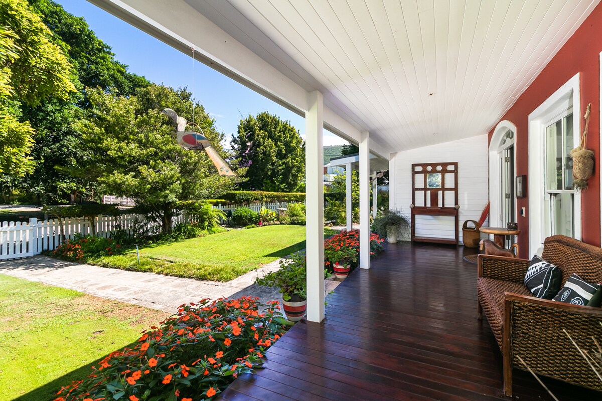 A charming porch features dark wooden flooring and is bordered by vibrant flower beds. A comfortable wicker seating area is situated to one side, while a nearby table reflects natural light. The surrounding greenery creates a peaceful outdoor setting, inviting relaxation.