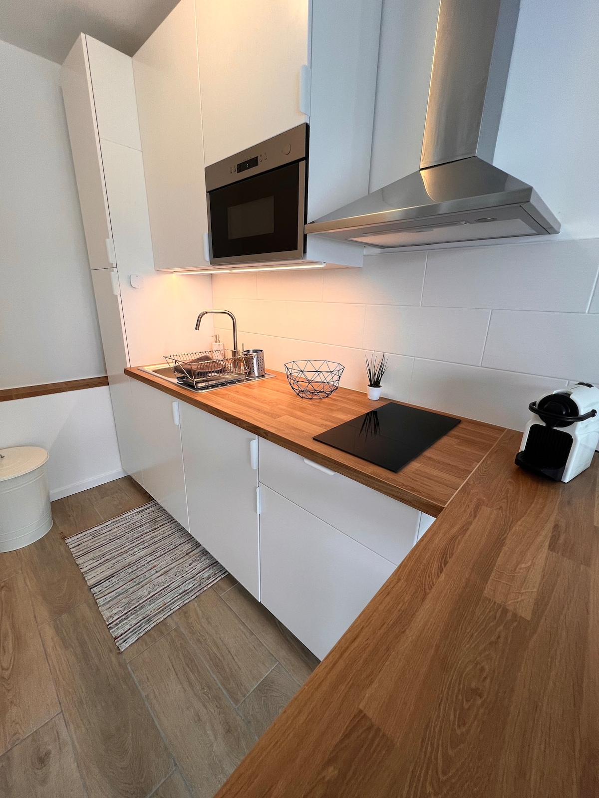 A modern kitchen space is presented, featuring a wooden countertop and sleek white cabinetry. Built-in appliances, including an oven and a coffee maker, are visible. A small decorative bowl and a plant add subtle accents to the area. A striped kitchen rug is placed on the floor.