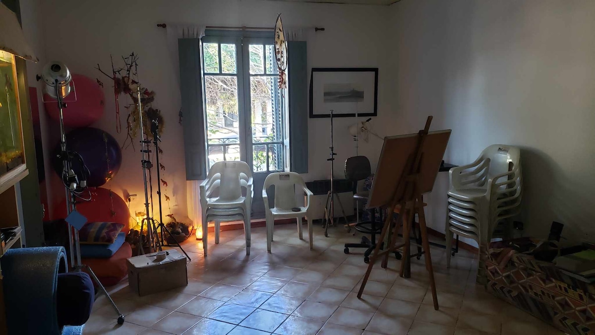 A cozy room features a set of stacked white plastic chairs and an art easel. Natural light enters through a window with sheer curtains, illuminating the space. Various art supplies and decorative items are arranged, creating a creative atmosphere for artistic expression.